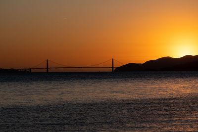 Silhouette bridge over sea against orange sky