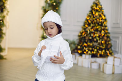 Boy in a white knitted sweater and hat stands with gift box  christmas tree at home on christmas day