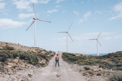 Rear view of man standing on dirt road amidst windmills against sky