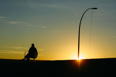 Rear view of silhouette man sitting on bench by street light against sunset sky