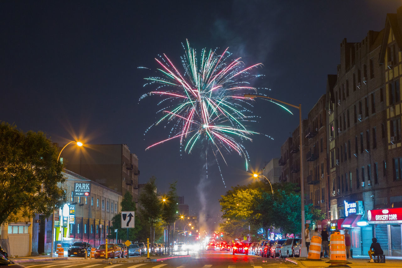 night, illuminated, firework display, celebration, blurred motion, motion, exploding, long exposure, firework - man made object, city, building exterior, architecture, arts culture and entertainment, street, outdoors, sky, city life, multi colored, low angle view, built structure, cityscape, firework, no people