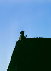 Low angle view of silhouette person on rock against clear blue sky