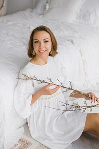 Young woman in a white dress holds an easter branch in her hands and sits at home by the bed