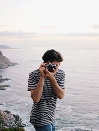 Young man photographing at beach against sky during sunset