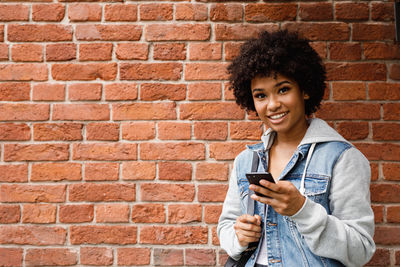 Portrait of smiling young man using mobile phone against brick wall
