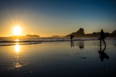 Silhouette people with surfboard walking on shore at beach against sky during sunset