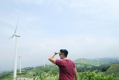 Man shielding eyes while looking away against windmills and sky on field