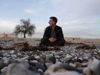 Mid adult man yawning while sitting on pebbles against sky