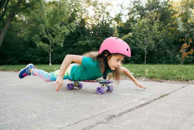 Happy smiling caucasian girl in pink helmet lying on tummy riding skateboard on road in park 