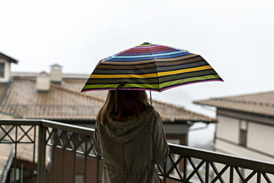 Young woman from behind with umbrella in rainbow pattern in drops standing on terrace in rain