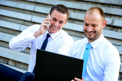 Happy businessman using phone while sitting by colleague with laptop