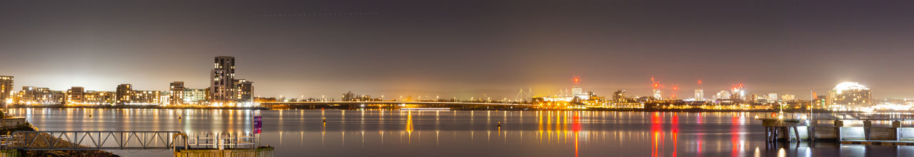 Illuminated buildings by river against sky at night