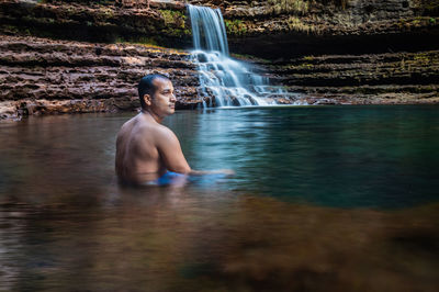 Young man sitting in natural waterfall clear water at morning from flat angle