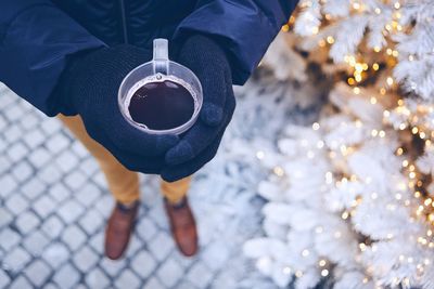 Low section of man holding black coffee while standing by illuminated christmas tree during winter