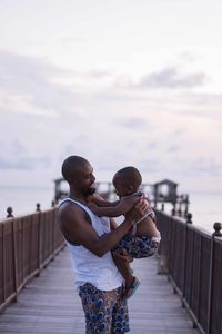 Father carrying son while standing on pier against sky during sunset