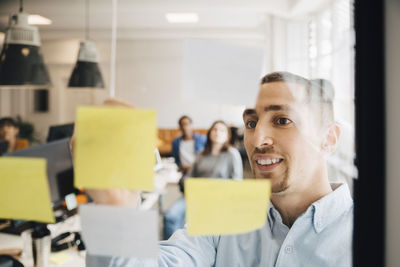 Male computer programmer sticking adhesive note on glass during meeting with colleagues in creative office