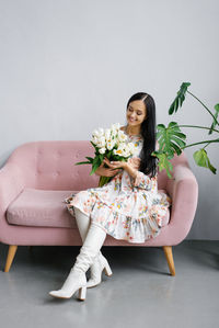 A young pretty happy woman in a dress and white boots sits on a pink sofa with a bouquet