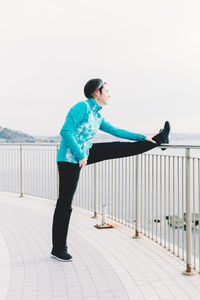 Side view of young man standing on railing against sky