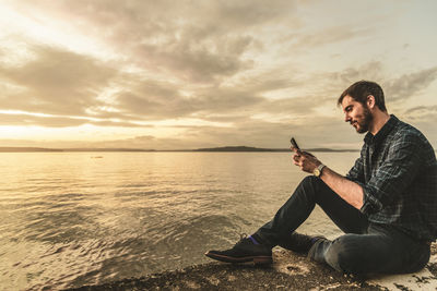 Man using mobile phone while sitting at sea against sky during sunset