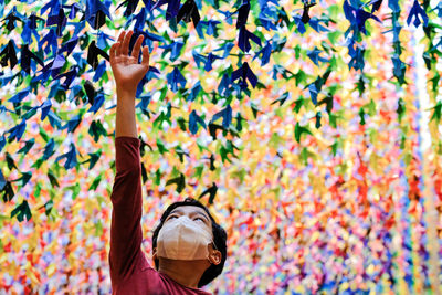 A boy with a face mask reaching up to the suspended origami birds