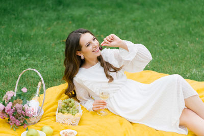 Beautiful young woman in a white dress drinking wine in the garden on a picnic