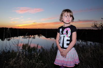 Girl looking away while standing at lakeshore against sky during sunset