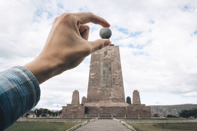 Optical illusion of cropped hand holding sphere shaped structure on building against cloudy sky