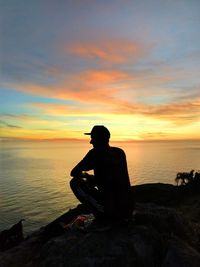 Silhouette man sitting on rock by sea against sky during sunset