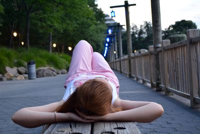 Young woman with hands behind head lying on bench against trees during sunset