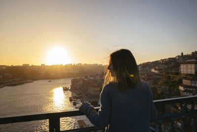 Rear view of woman looking at cityscape against sky during sunset