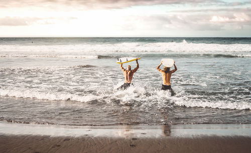 Rear view of men holding surfboard walking at shore against sky