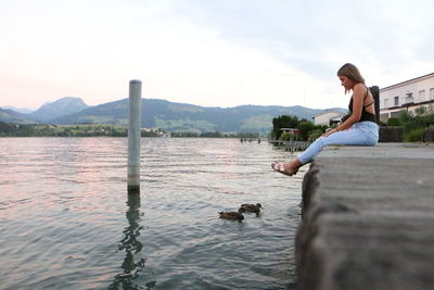 Full length of woman sitting on pier over ducks swimming on lake against sky