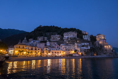 Illuminated buildings by sea against clear sky at night