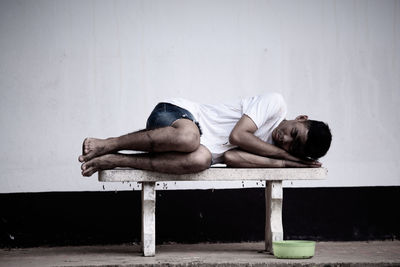 Portrait of young man sitting on bench against wall