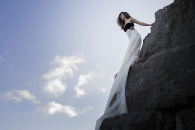 Low angle view of woman in evening gown standing on rock against sky