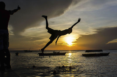 Silhouette man jumping in river against cloudy sky during sunset
