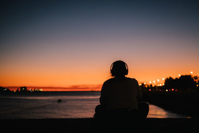 Silhouette woman wearing headphones by sea sitting against orange sky