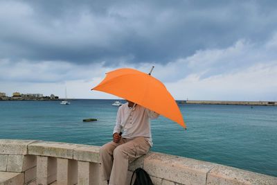 Woman sitting on umbrella by sea against sky