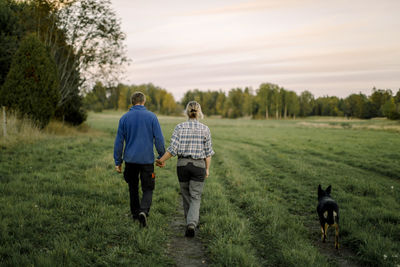 Couple holding hands and walking with dog on field at sunset