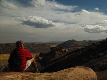 Rear view of man sitting on rock formation against mountains
