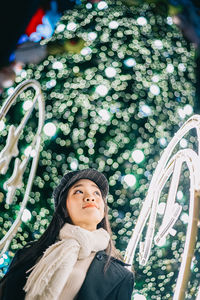 Low angle view of young woman looking up against illuminated christmas tree at night