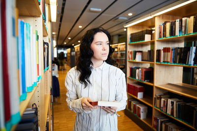 Young female student holding book while standing in university library