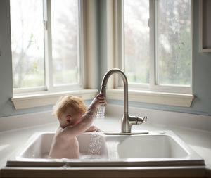 Cute baby boy holding faucet while sitting in kitchen sink against windows at home