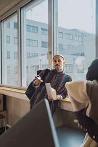 Portrait of confident male student with smart phone and book leaning on window sill in classroom