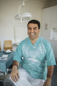 Portrait of smiling male surgeon wearing protective sitting in medical examination room
