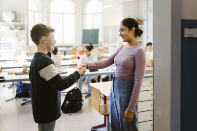 Smiling teacher shaking hand with schoolboy standing in classroom