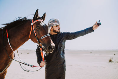 Young man taking selfie with mobile phone riding horse at desert