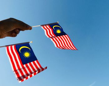 Low angle view of hand holding malaysian flag against clear blue sky