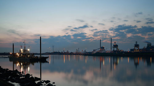 Harbor in river by industrial district against cloudy sky during sunset