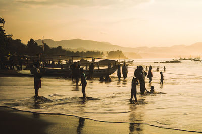 People on shore at beach against sky during sunset